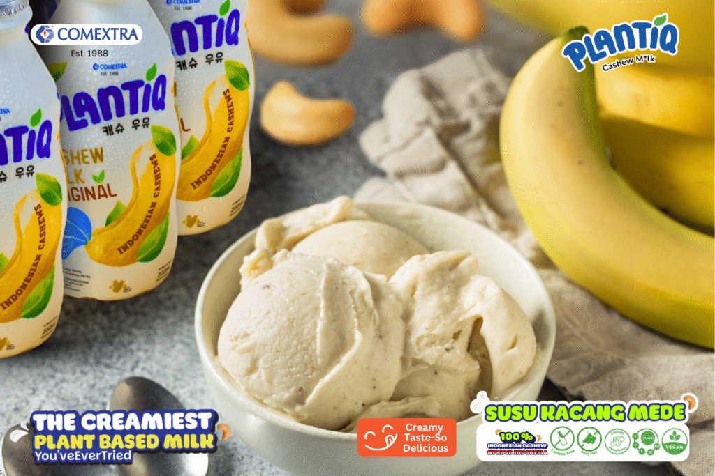 Semangkuk es krim dengan susu PLANTIQ original di sisi kiri, setandan pisang, kacang mede, dan sepasang sendok di kiri bawah. 
